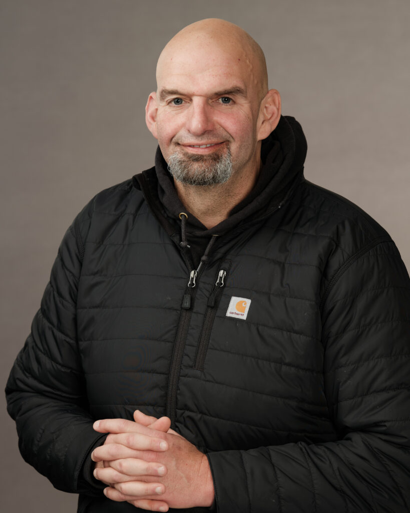 A photo of John Fetterman, smiling, with hands clasped. He is wearing a black Carhartt sweater jacket.